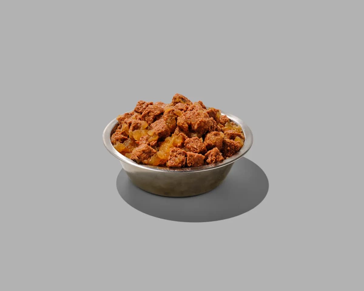 A metal bowl filled with chunks of fresh dog food on a gray background.
