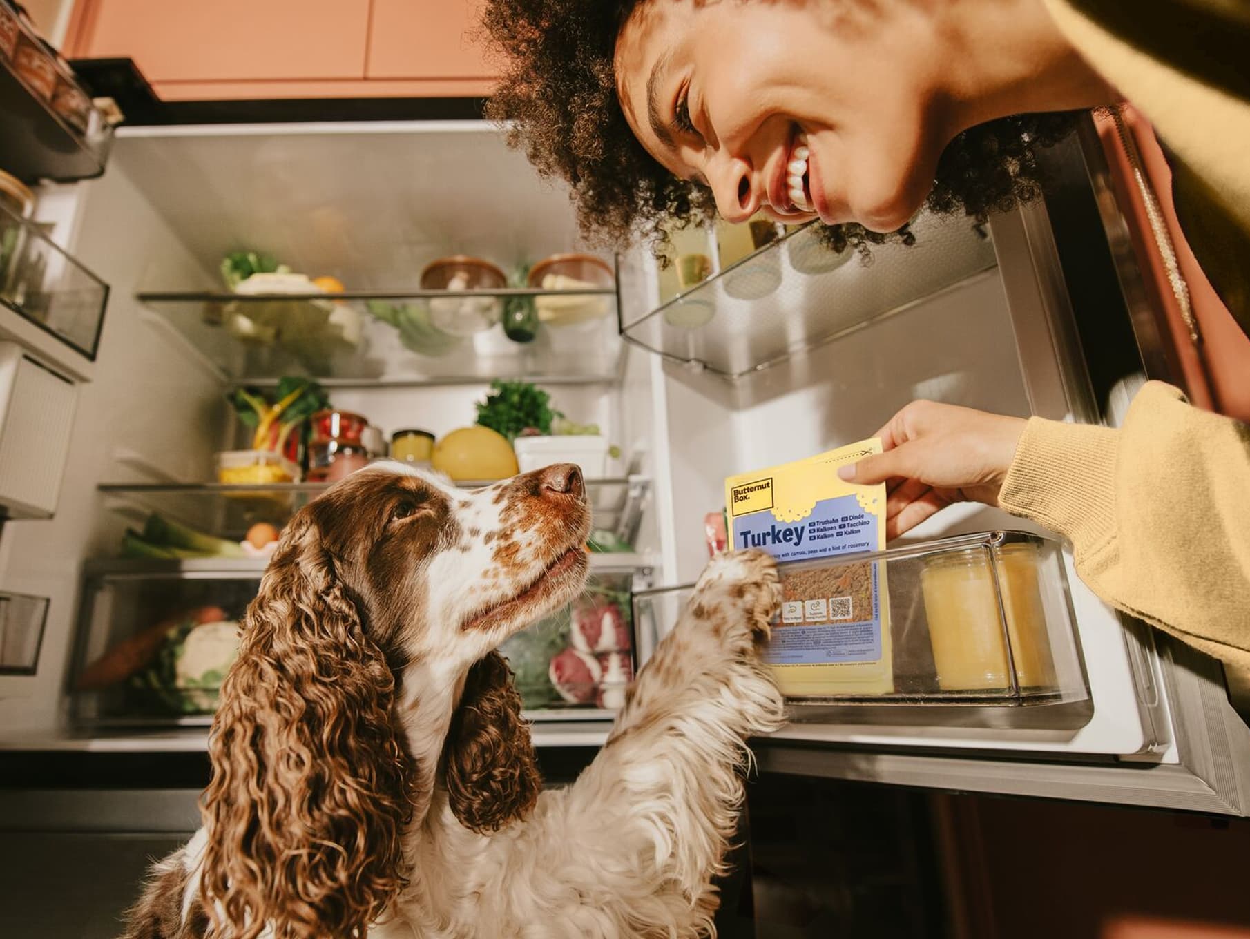 A woman smiles at her dog as she retrieves a pack of Butternut Box Turkey dog food from the refrigerator.