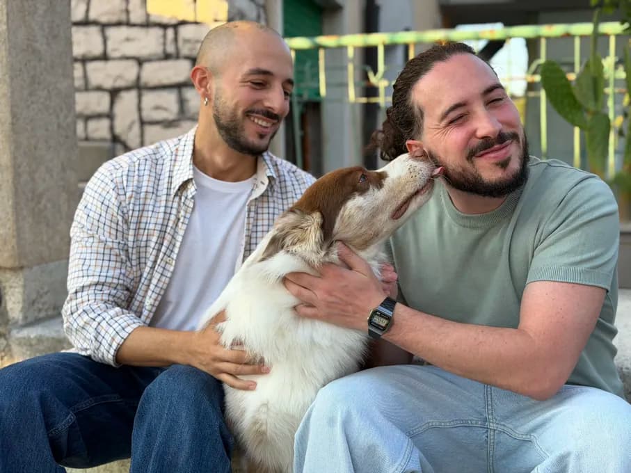 Two men sitting on steps, smiling as they pet a dog. The dog is playfully licking one man's face.