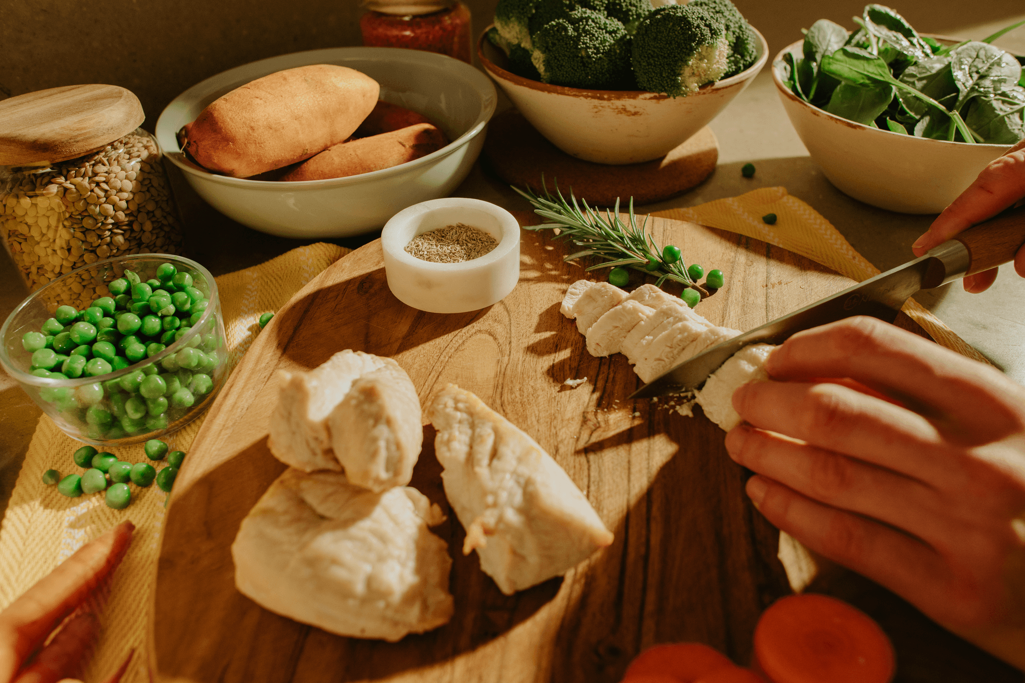 Hand slicing cooked chicken on a wooden board, surrounded by peas, sweet potatoes, rosemary and bowls of greens.